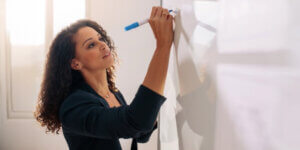 A woman standing in front of a whiteboard, holding a marker and writing on it with focused concentration.