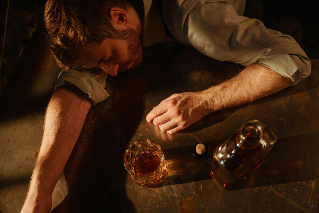 a man slumped over a table with a whisky bottle and glass in front of him