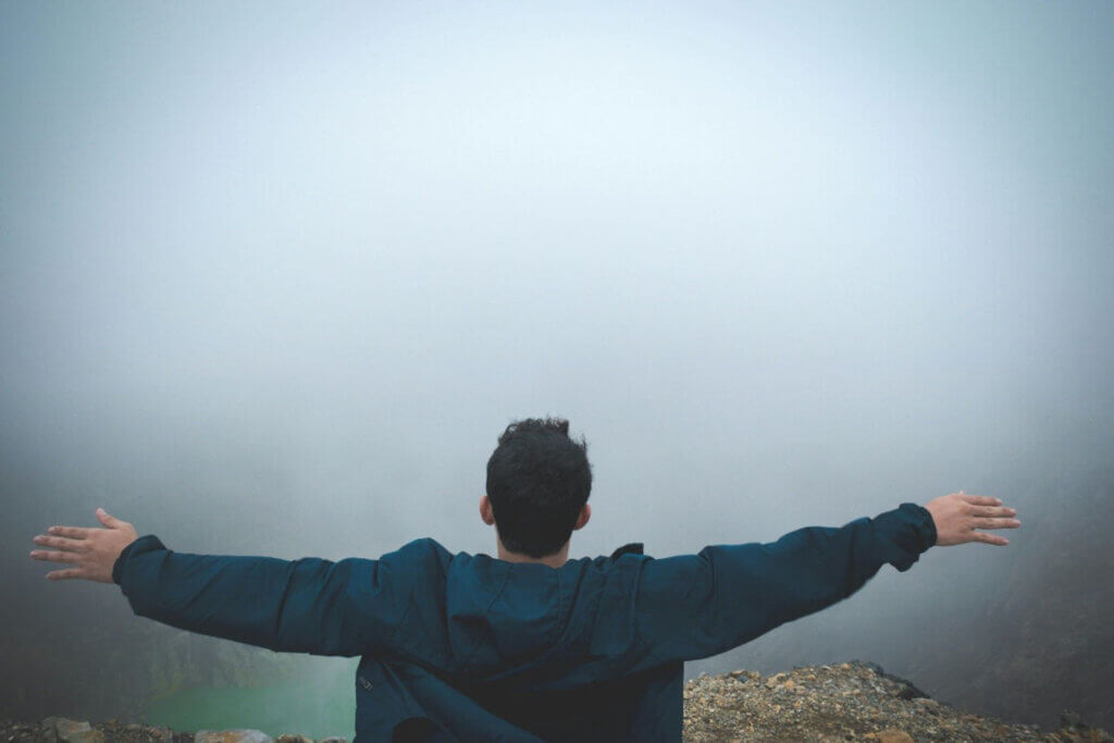 a man stands at the edge of a cliff with arms outstretched looking into the mist