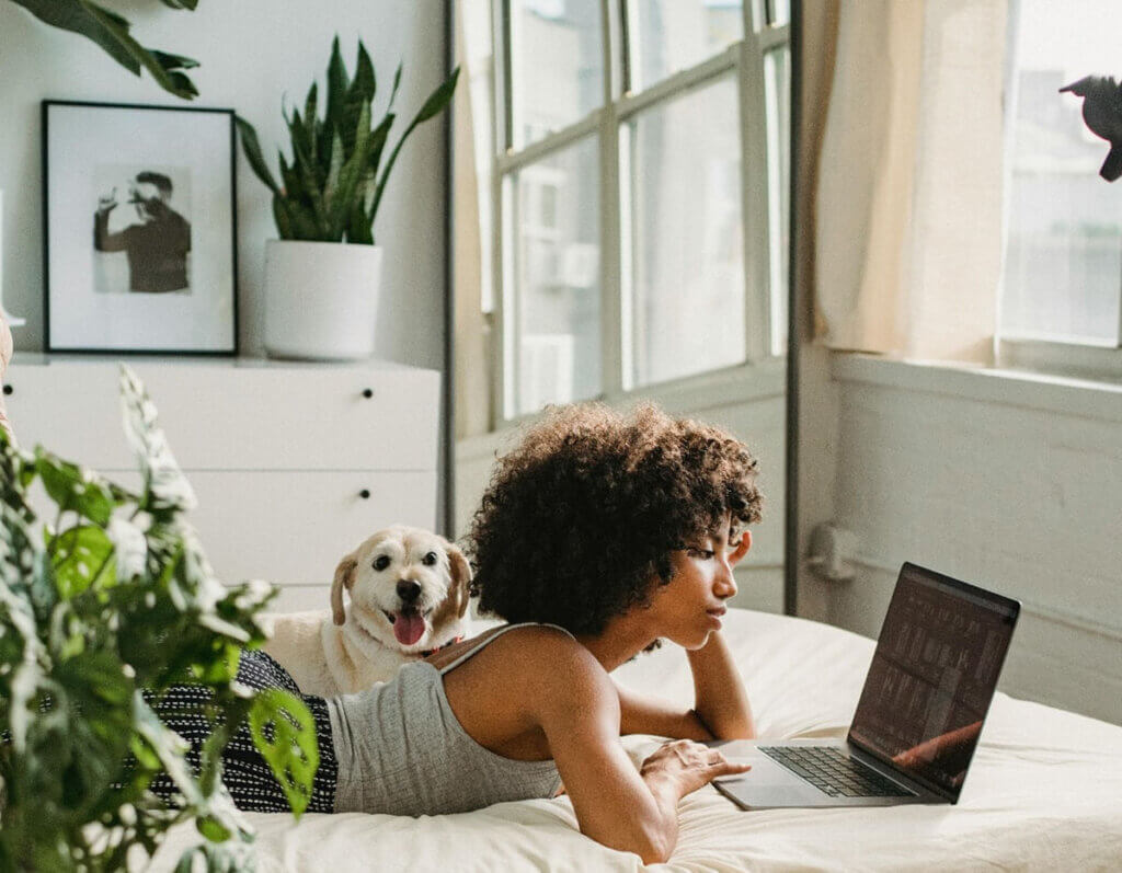 a woman lays on a bed with her dog surrounded by plants and looks at her computer