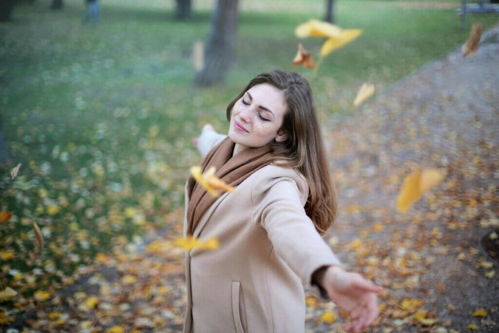 woman standing with her arms outstretched and her eyes closed standing in falling autumn leaves