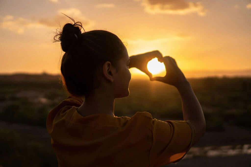 A woman making a heart with her hands at sunset.