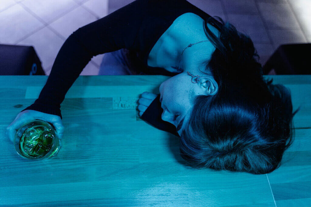 A woman sleeping while leaning on a table, with a glass of alcohol in her hand.