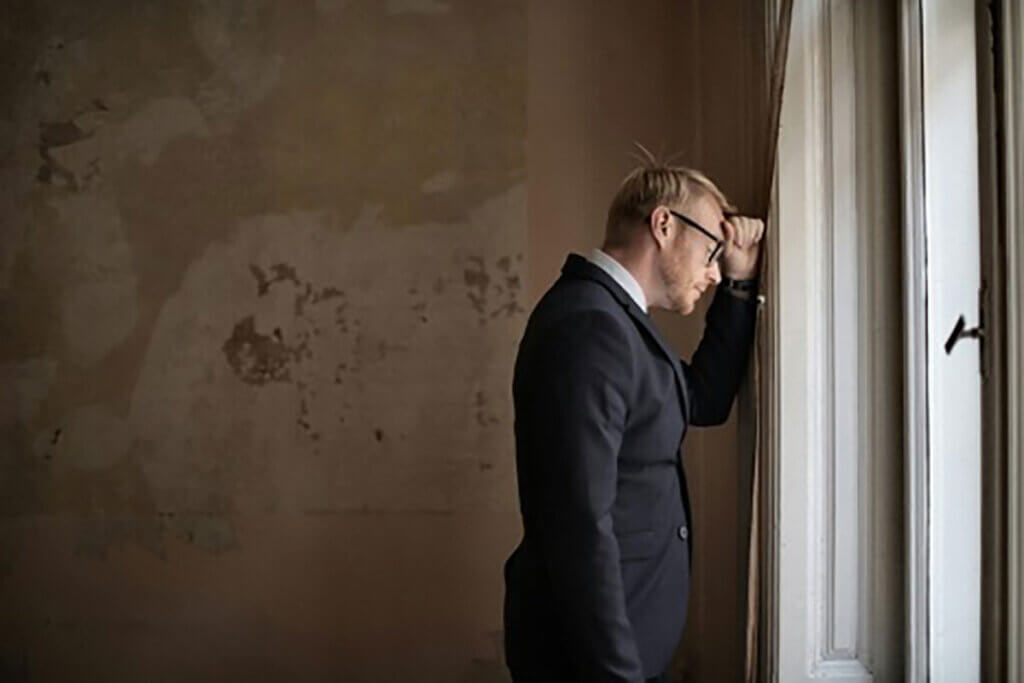 A man leaning against a window while looking distressed.