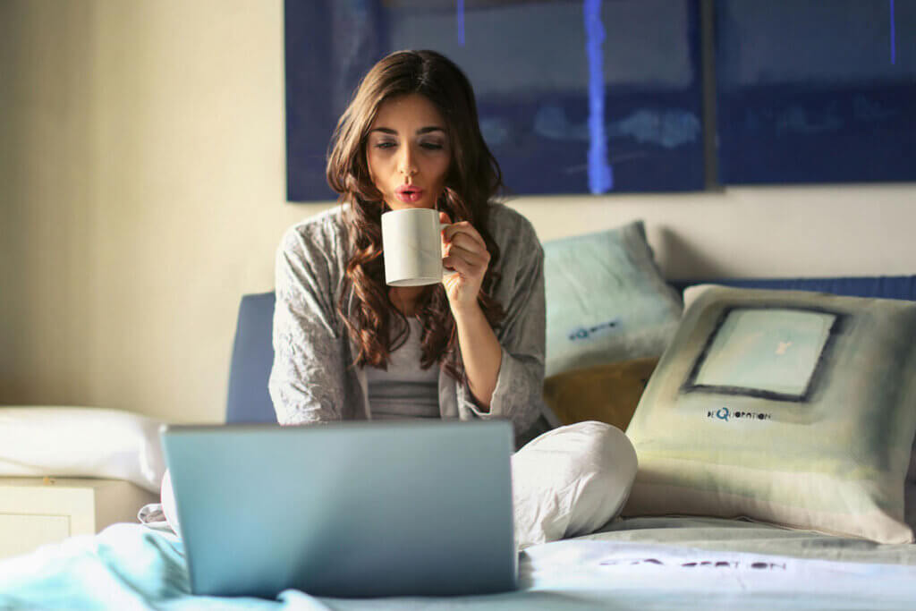 A woman sitting on a bed drinking coffee and looking at a laptop.