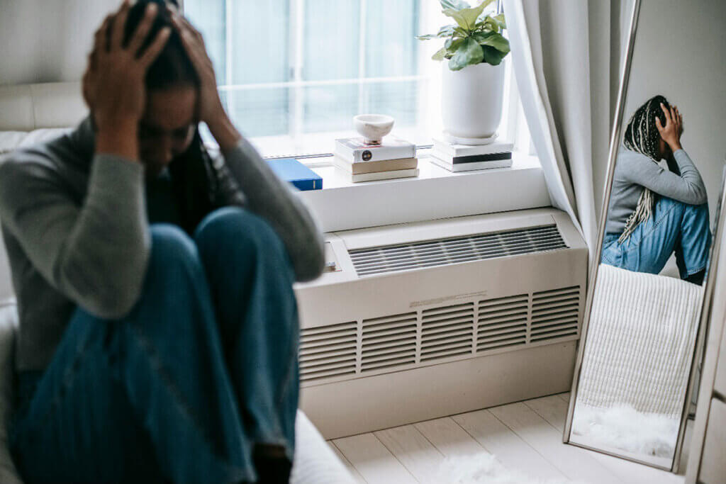 A woman sitting on a couch holding her head.. She looks distressed and her reflection can be seen in a mirror.