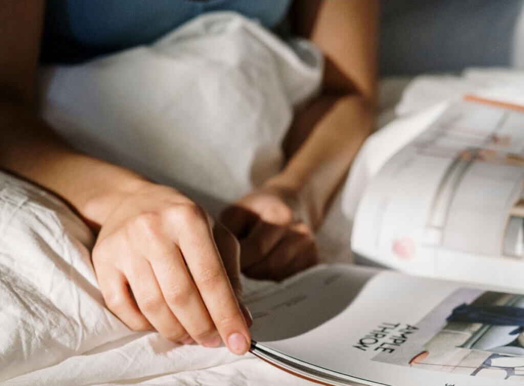 A person's hands and arms and a book on a bed.