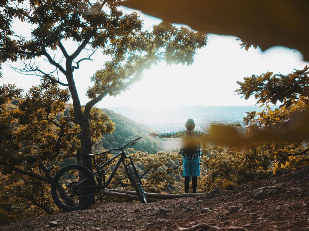 A person standing next to a bicycle in nature with their arms stretched out.