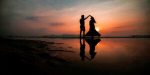Silhouette of a couple dancing by the water at sunset, with their reflection shimmering on the wet sand.