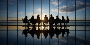 Silhouetted business professionals sit around a conference table in a high-rise office with floor-to-ceiling windows, as the sun sets over a calm ocean horizon, reflecting off the polished floor.