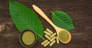 Green kratom leaves, powder in a spoon and bowl, and capsules are arranged on a wooden surface.