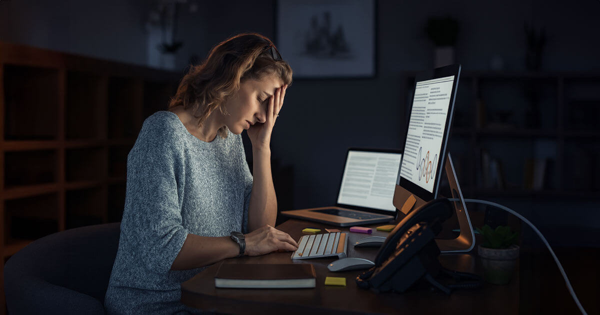 Stressed woman sits at a desk late at night with her hand on her forehead, looking exhausted in front of a computer screen.