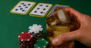Hand holding a glass of whiskey with ice next to poker chips and playing cards on a green table.